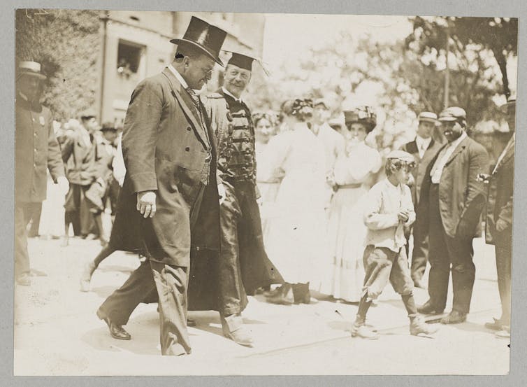 A sepia-toned photograph of two men in formal clothing and academic robes walking and talking.