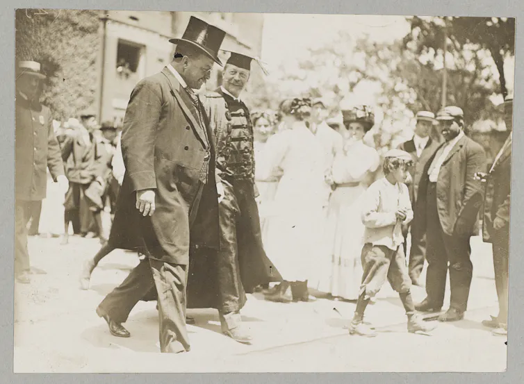 A sepia-toned photograph of two men in formal clothing and academic robes walking and talking.