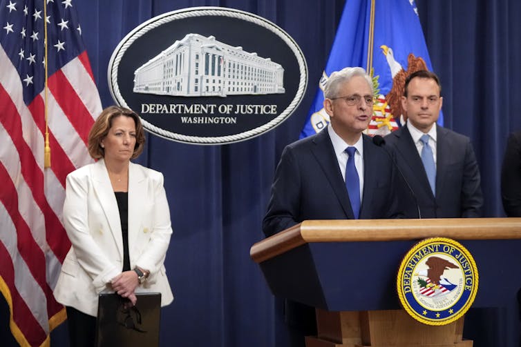 two men and a woman in business attire stand behind a lectern and in front of flags and a logo