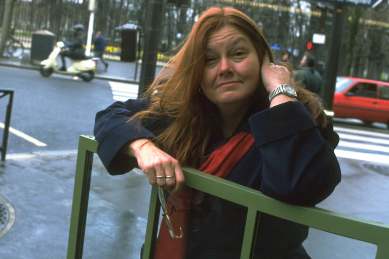 Middle aged woman with long red hair stands on a sidewalk holding a pair of eyeglasses.