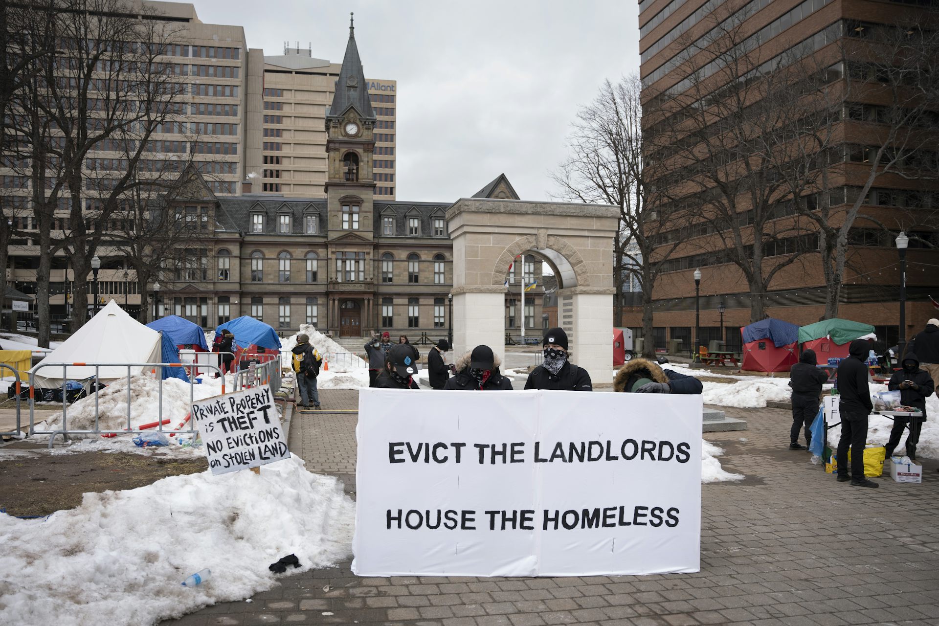 Protesters hold up a sign reading Evict the Landlords, House the Homeless at a snowy homeless camp.