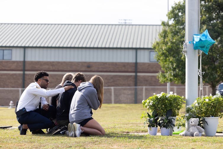 Three students kneel at a memorial as a chaplain comforts them from behind.