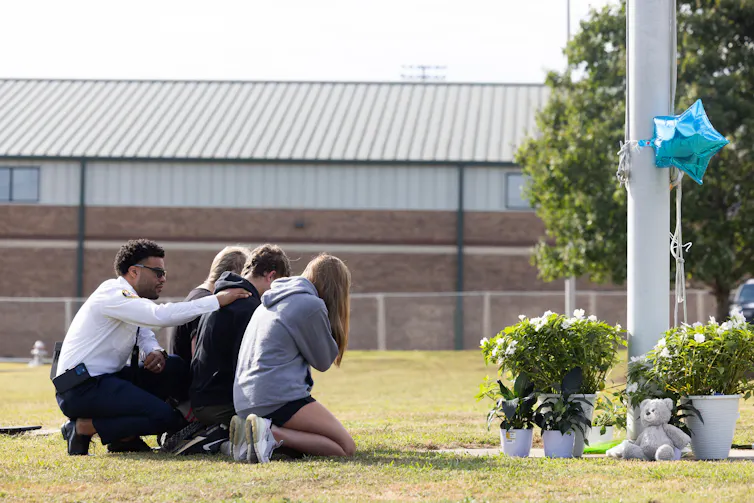 Three students kneel at a memorial as a chaplain comforts them from behind.