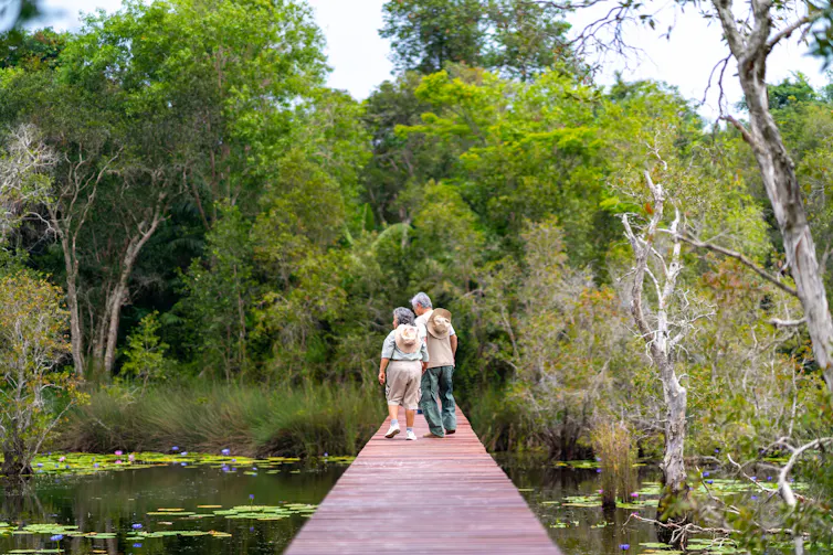 An older Asian couple walk on a wooden bridge out in nature.