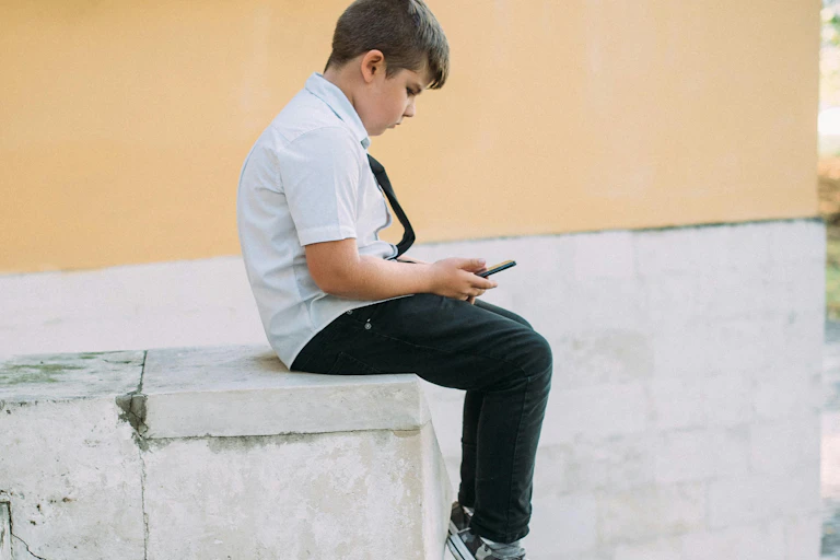 A boy in a white shirt and black tie sits alone looking at his phone.