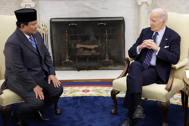 One man in a traditional Indonesian hat sits on a chair opposite a man in a suit.