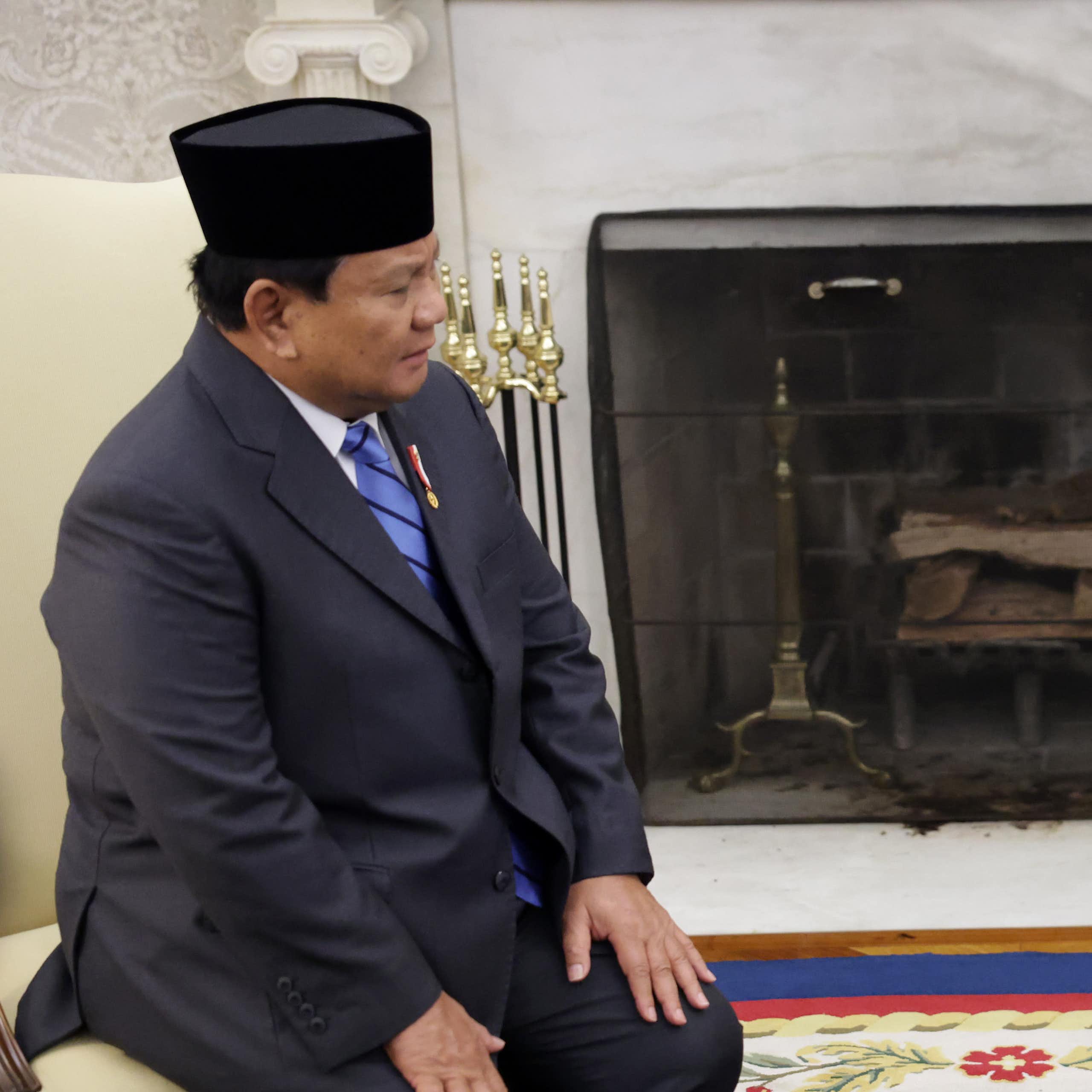One man in a traditional Indonesian hat sits on a chair opposite a man in a suit.