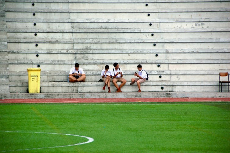 A distant photo of four boys in school uniforms sitting in a stadium and looking at their phones.