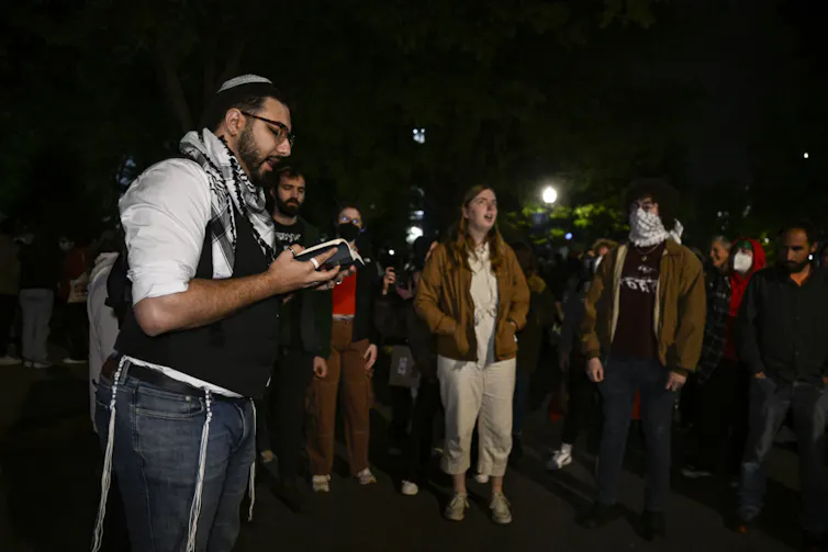 Young people in light jackets stand outside at nighttime as one young man reads aloud from a book.