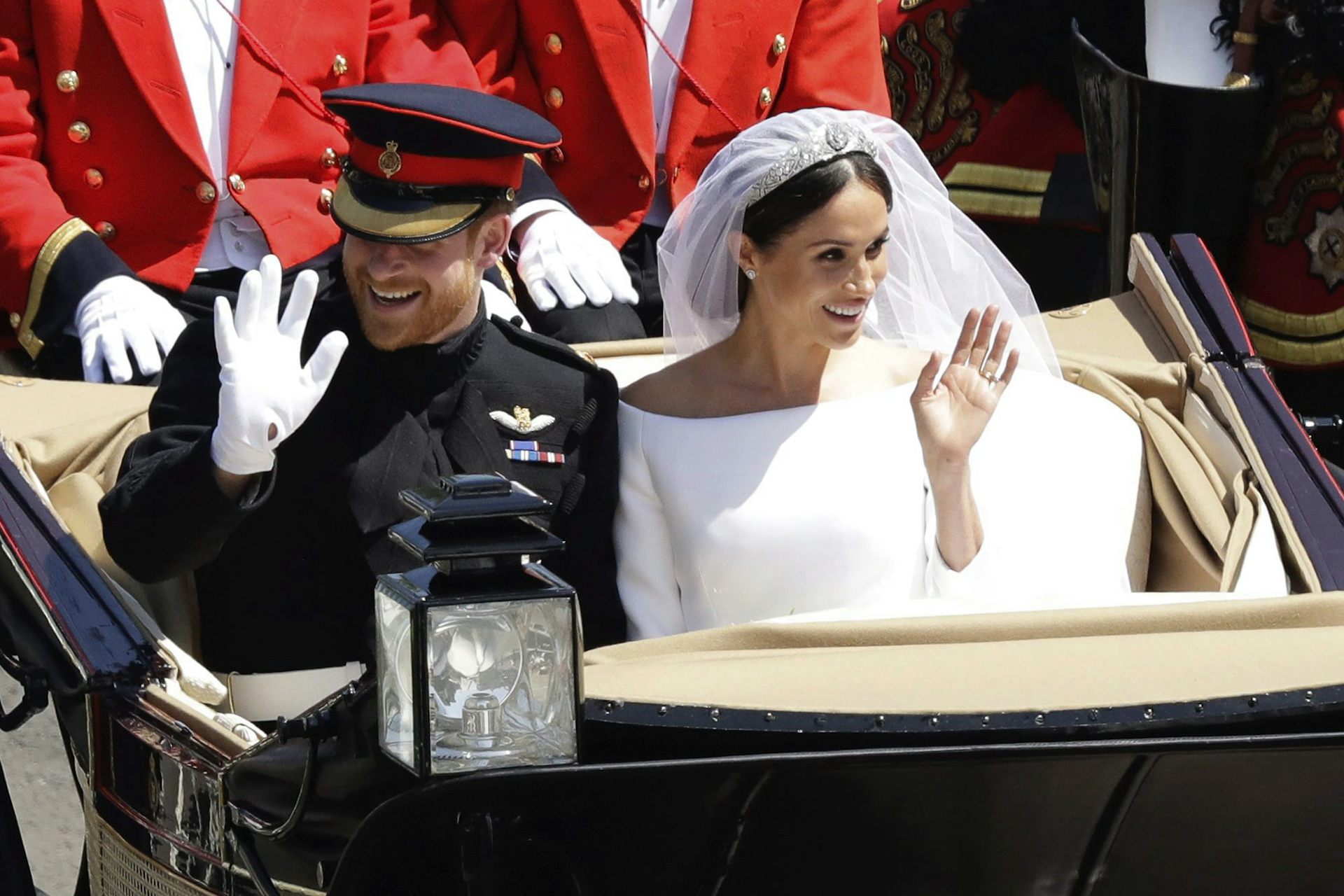 A dark-haired woman in a white wedding gown and veil smiles and waves from a horse-drawn carriage. Her red-haired and bearded husband sits beside her smiling and waving.