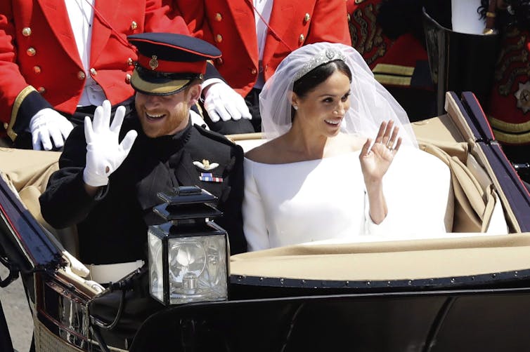 A dark-haired woman in a white wedding gown and veil smiles and waves from a horse-drawn carriage. Her red-haired and bearded husband sits beside her smiling and waving.