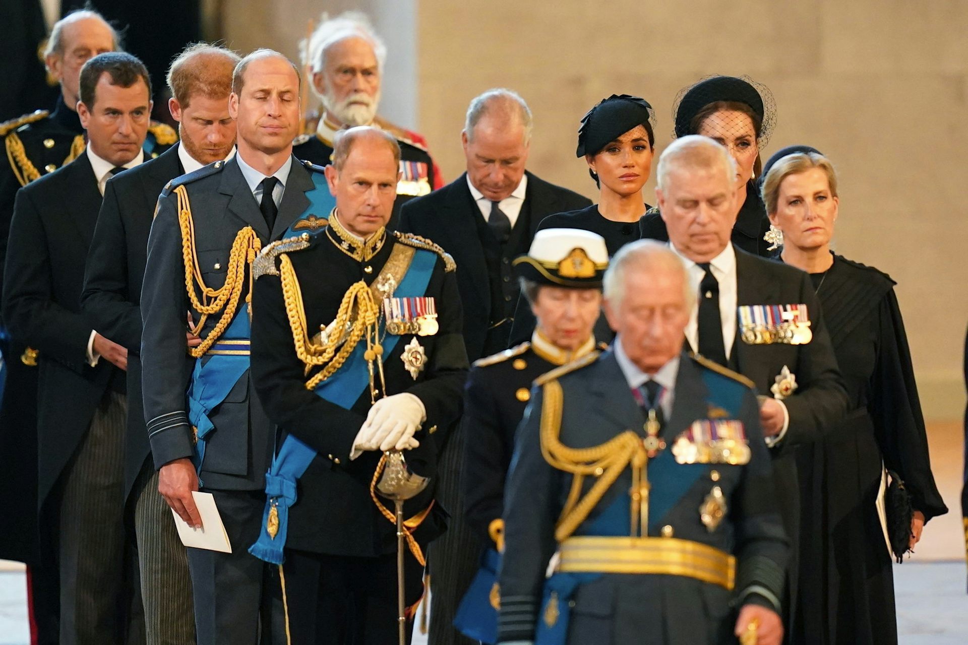 A sole Black woman walks among a group of white people in military uniforms or black suits and dresses.