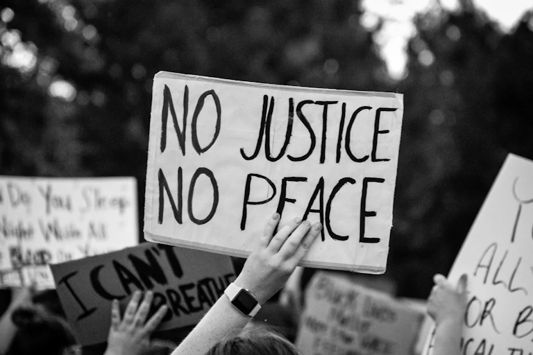A black-and-white photo shows a protester holding up a No Justice, No Peace sign.
