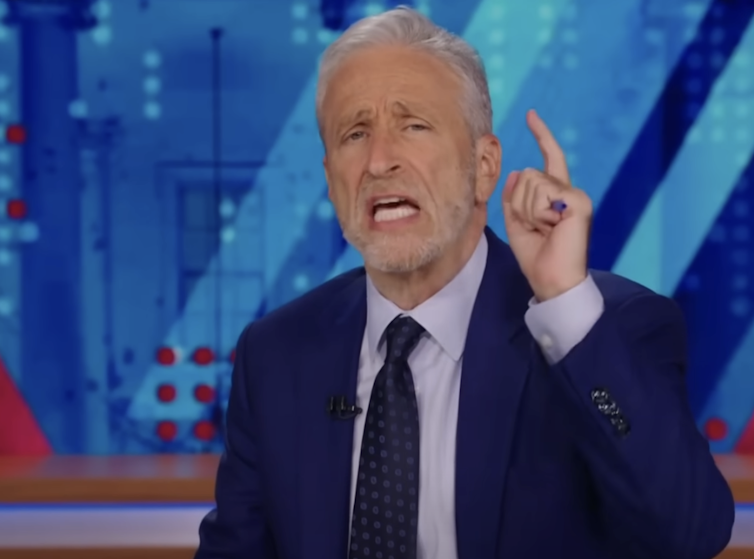 A man in a dark blue blazer speaking and raising his left hand to make a point.