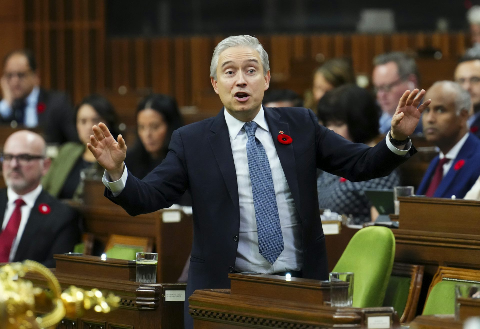 A grey-haired man in a blue suit with a red poppy on the lapel gestures as he speaks in the House of Commons.