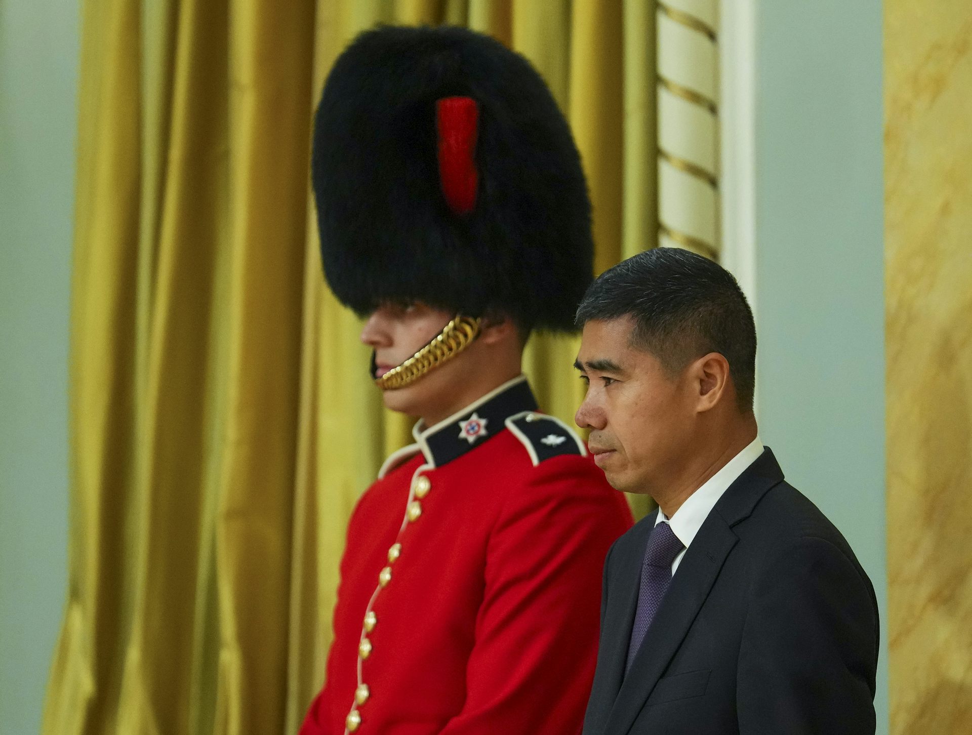 An Asian man stands beside a ceremonial military guard in a red jacket and a tall black fur hat.