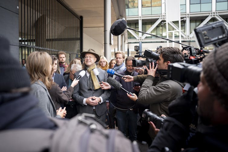 A person mobbed by reporters outside a courtroom.