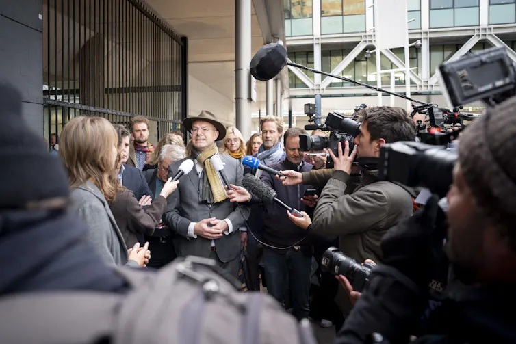 A person mobbed by reporters outside a courtroom.