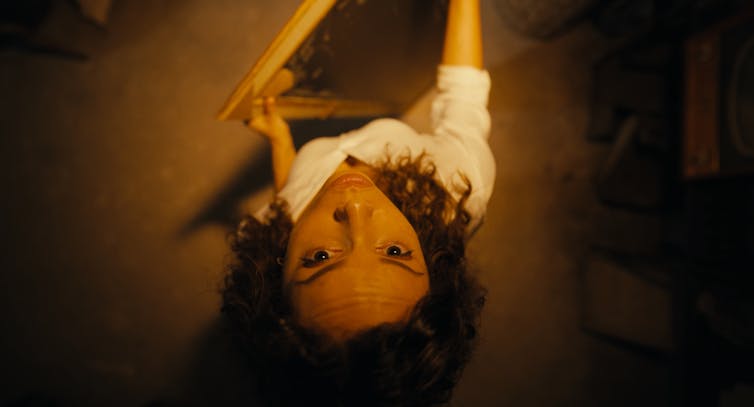 A brunette woman in a white shirt looks up toward the camera, facing the top of the frame.