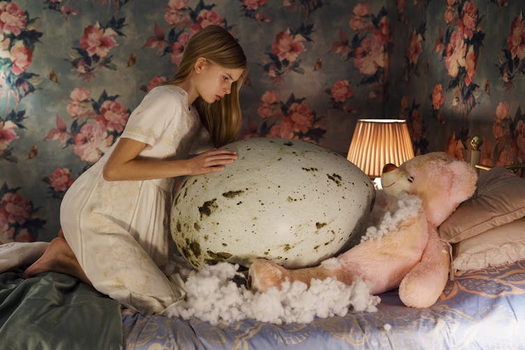A girl in a white dress crouches on a bed with a large pink teddy bear and a huge bird egg as large as she is.