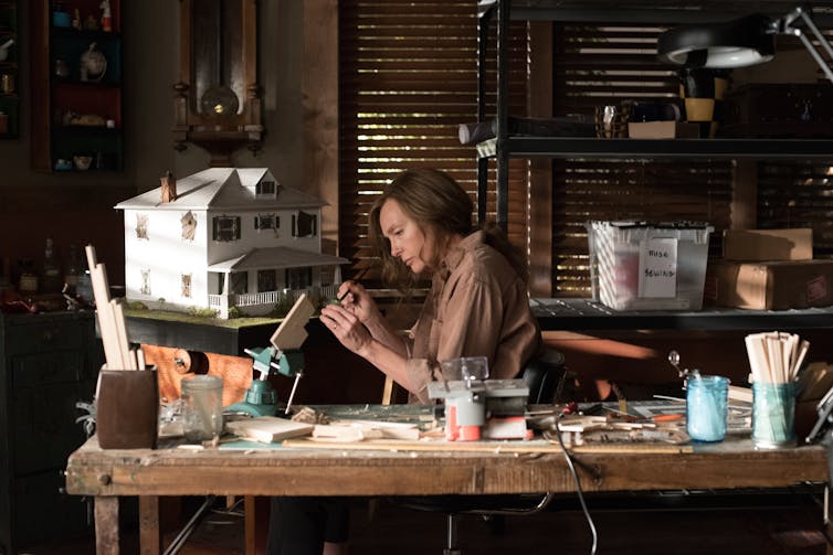 A woman works intently on a small craft, sitting in a cluttered workshop in front of a white dollhouse.