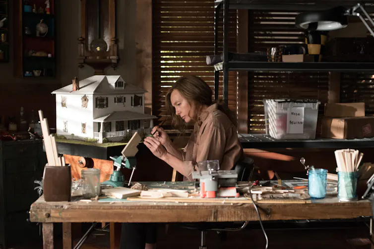 A woman works intently on a small craft, sitting in a cluttered workshop in front of a white dollhouse.