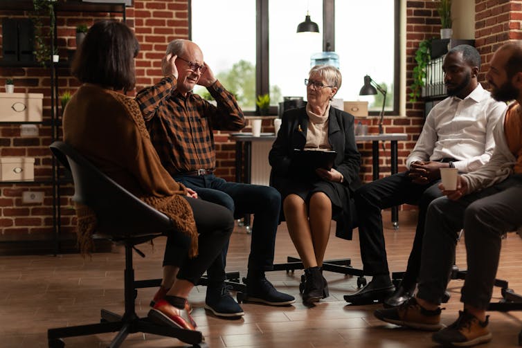 Five people sitting in a circle. One is speaking and holding his hands to his head.