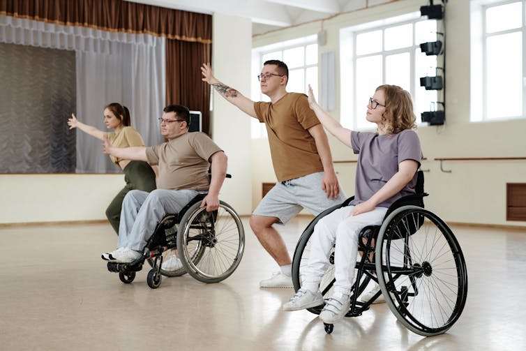 Four people in a dance studio, two of whom are using wheelchairs, with their arm stretched to the right