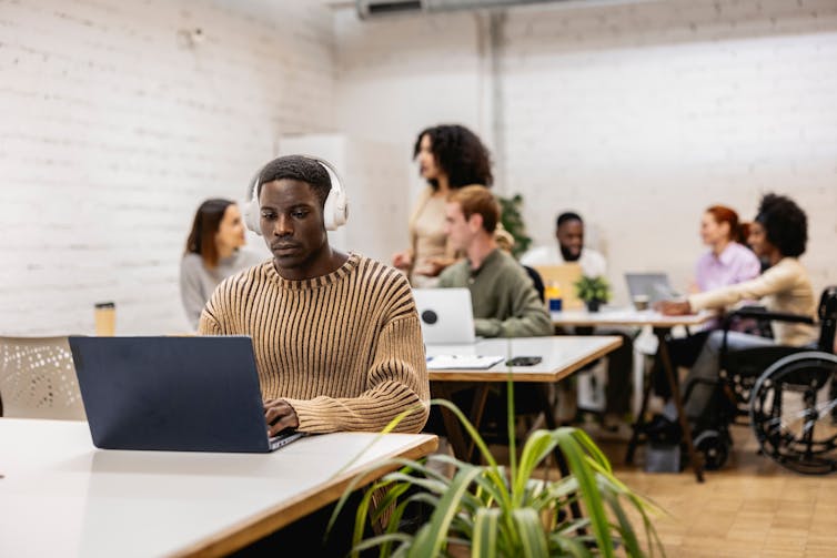 Busy office with young man in foreground at desk wearing headphones and people interacting the background