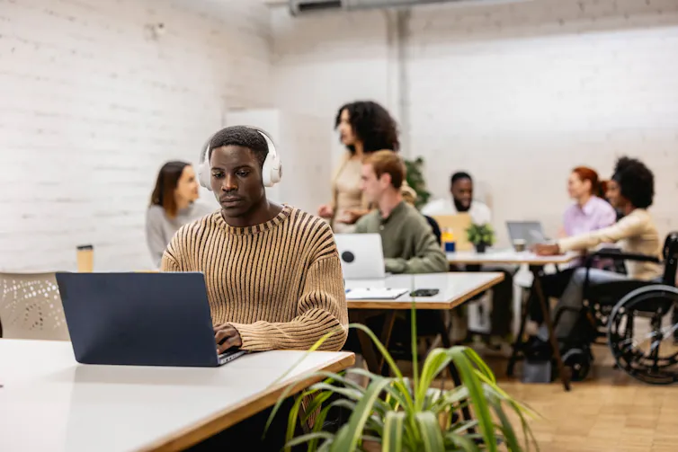Busy office with young man in foreground at desk wearing headphones and people interacting the background