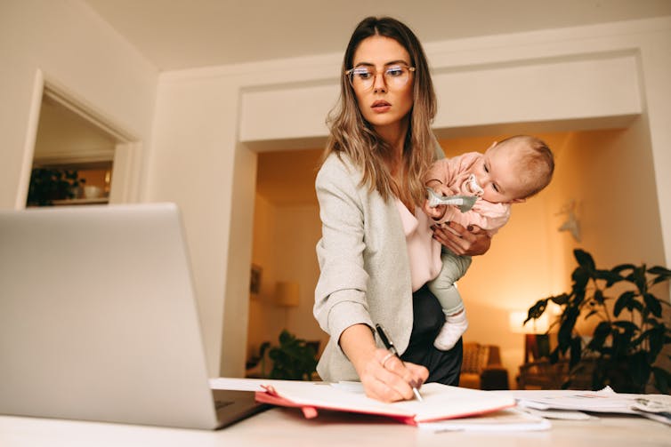 A woman writes in a notebook while looking at a laptop sitting open on a desk and holding a child in one arm