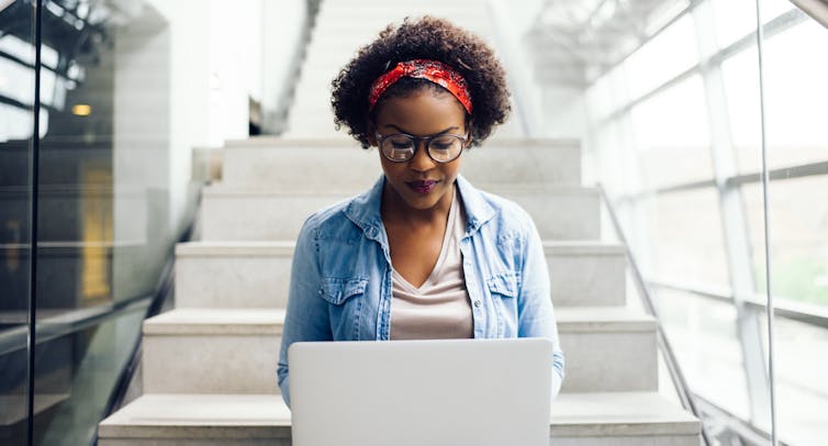 A young woman works on a laptop while sitting on a set of stairs