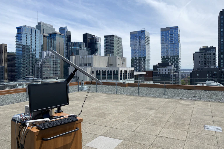 A computer attached to a solar panel on a rooftop, tall glass buildings in the background