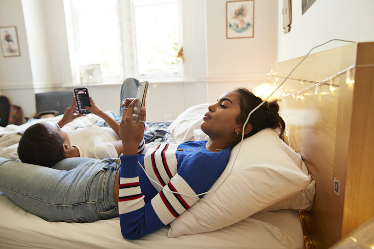 a young woman lays in bed looking at her phone while a young man lays against her thigh while looking at his phone