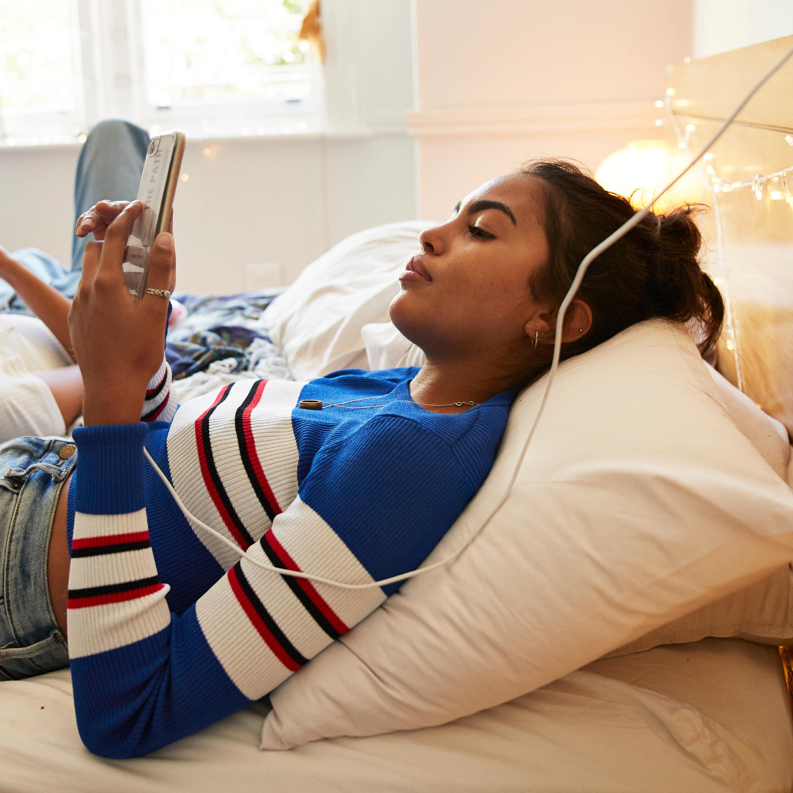 a young woman lays in bed looking at her phone while a young man lays against her thigh while looking at his phone