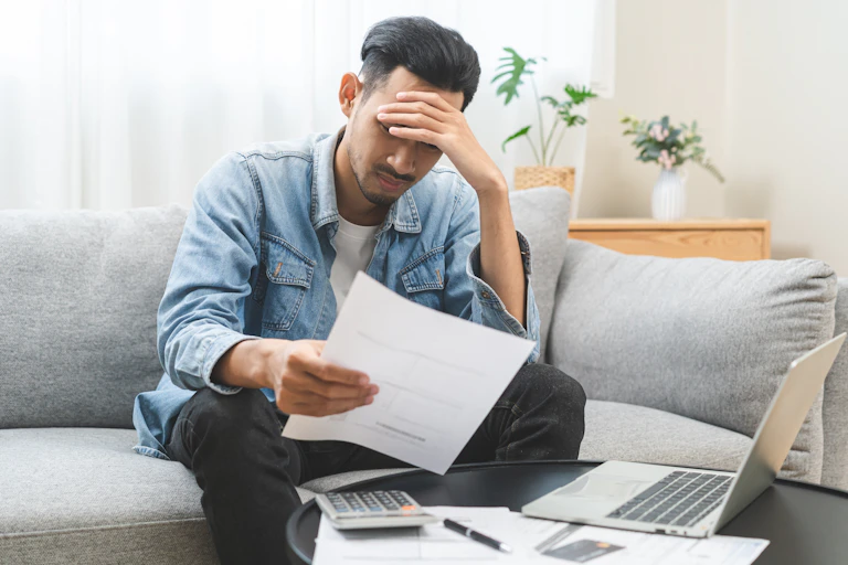 A man sits on a couch and rests his had against his hand while he looks at a piece of paper in his hand in concern