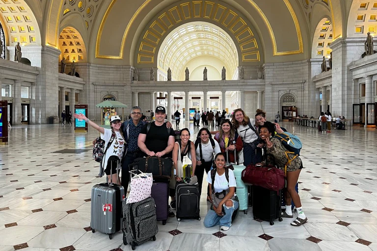 A group of young people smile and pose together in a large empty hall. They all have suitcases and backpacks with them.