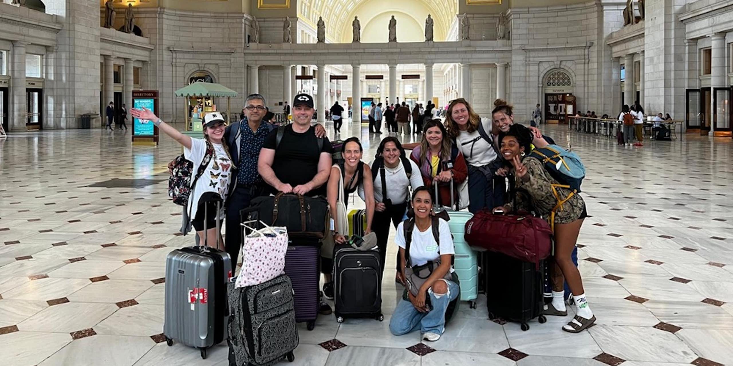 A group of young people smile and pose together in a large empty hall. They all have suitcases and backpacks with them.