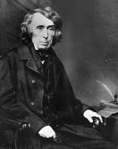 A vintage photo of a dark-haired man in dark clothes, sitting in a chair.