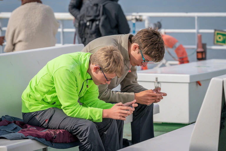 Two teenager boys on a ferry bent over, staring at their phones.