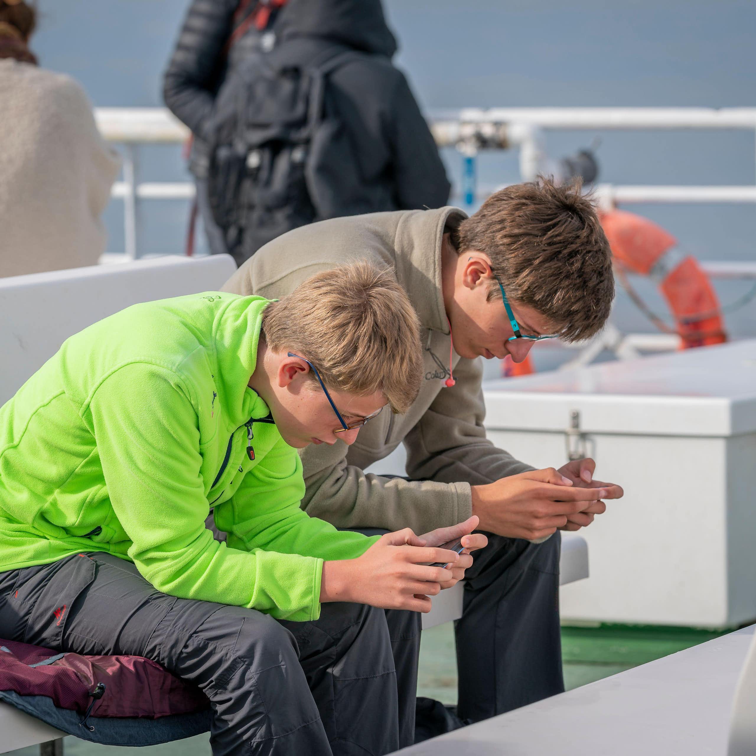 Two teenager boys on a ferry bent over, staring at their phones.