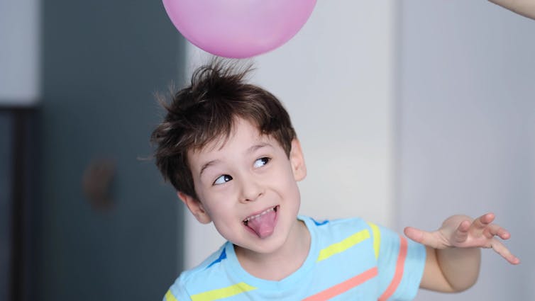 A child in a stripy shirt having a pink balloon rubbed on their head.
