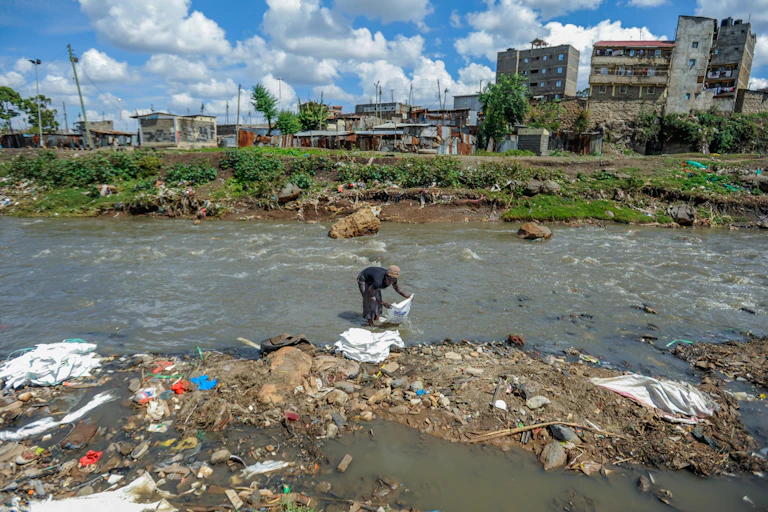 A person holding a big bag gathers litter from an urban river. There are homes and apartment blocks in the background