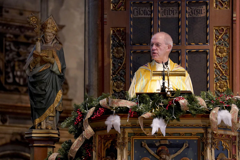 Justin Welby in a pulpit delivering a sermon next to a religious statue.