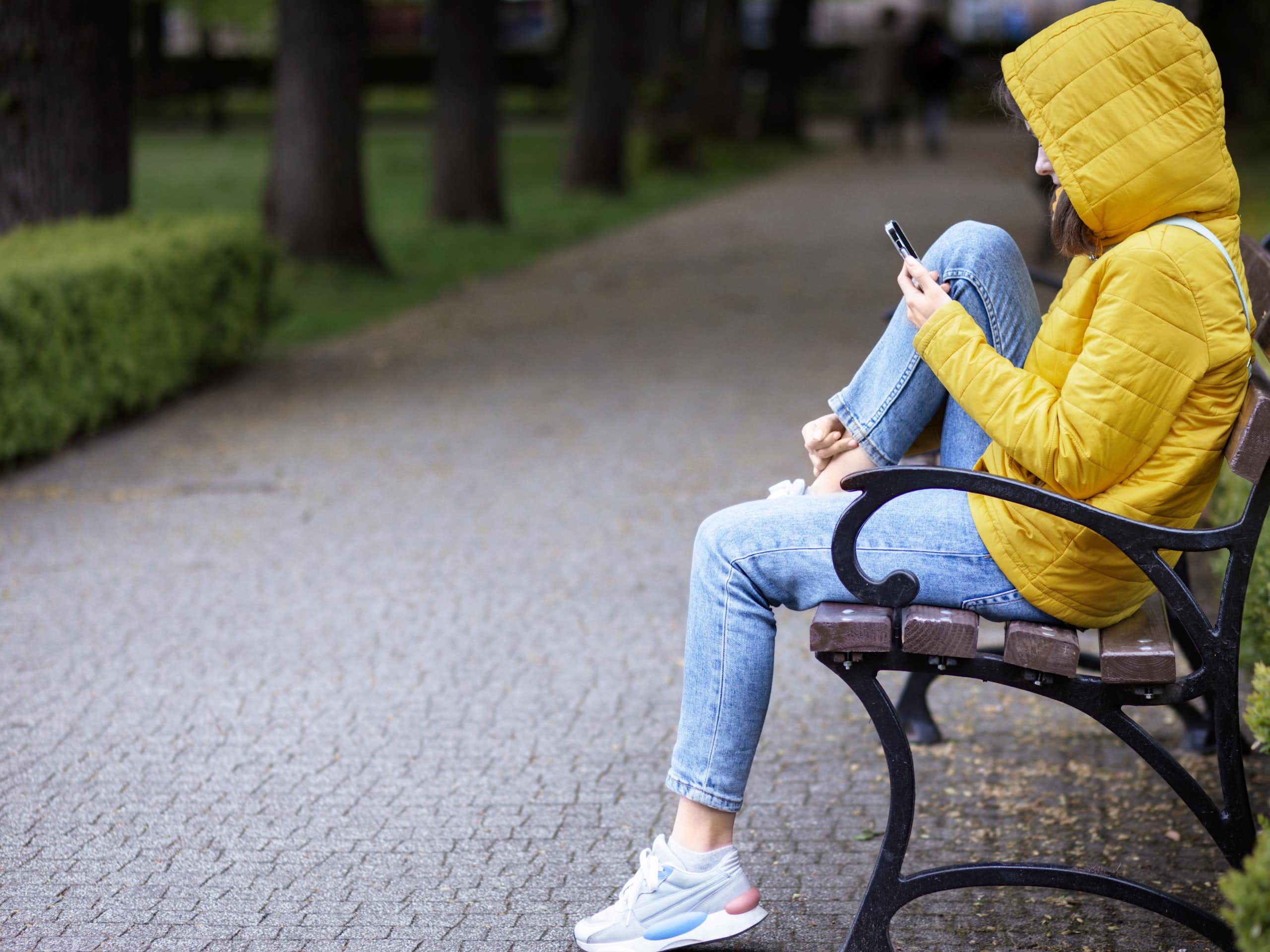 A young person in a yellow jacket is using a smartphone on a park bench.