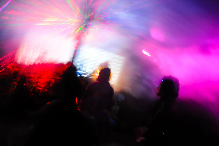 Image de nuit trippée avec des gens dansant dehors dans un festival de musique en forêt avec des poutres laser en arrière-plan.