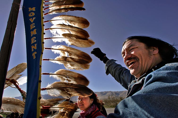 A man in a denim jacket raises his fist as he walks carrying a staff with feathers.