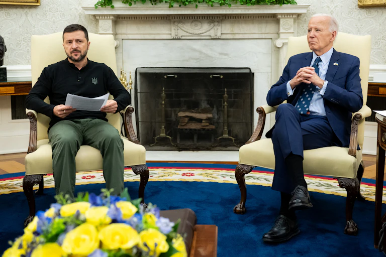 Two men, one in a suit, the other in a polo-neck shirt, sit on ornate chairs.