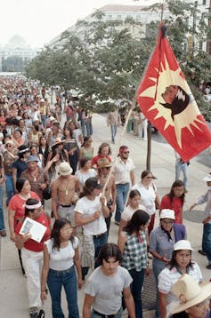 A faded, colored photo of a stream of people in summer clothing walking along a paved path, holding a flag.
