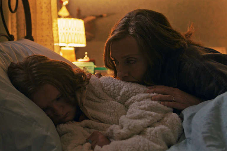 A woman looks intently at a girl sleeping in a bed, sitting beside it and resting her hand on the girl.